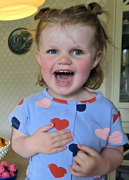 Ella 3 år har en ljusblå tröja med röda, rosa och blå hjärtan. Hon har två tofsar på huvudet och skrattar stort.