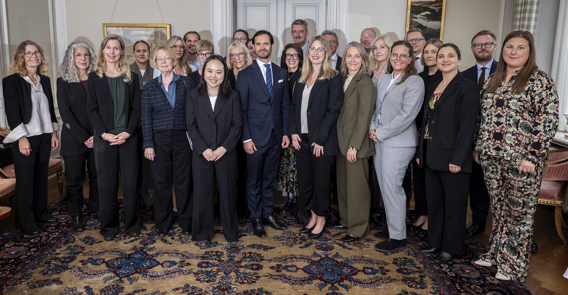 Ett foto med ett tjugotal personer som står på en tjock matta i beige och blått. I mitten står Prins Carl Philip med blå kavaj och slips.