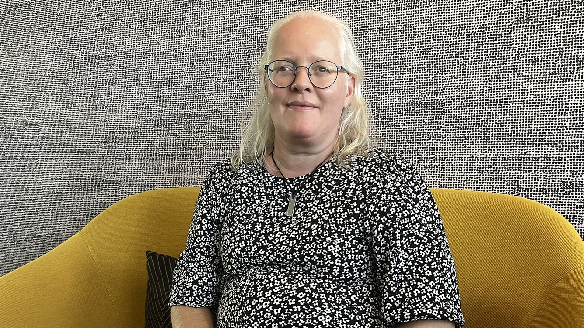 Ett foto av Linda Eriksson. Linda har glasögonen och en svartvit blus och sitter i en gul soffa.