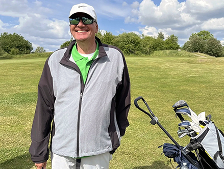 Lars står på en golfbana med golfbagen bredvid sig. Han har en tunn jacka, keps och solglasögon på sig.