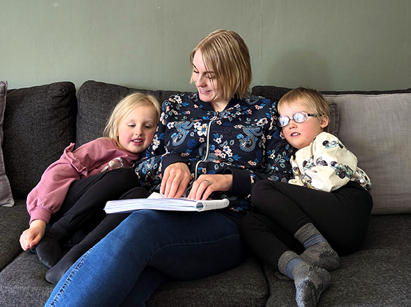 Maria sitter tillsammans med två av sina barn i en soffa. Hon läser i en punktskriftsbok. Barnen sitter på varsin sida om henne och lutar sig mot hennes axlar.