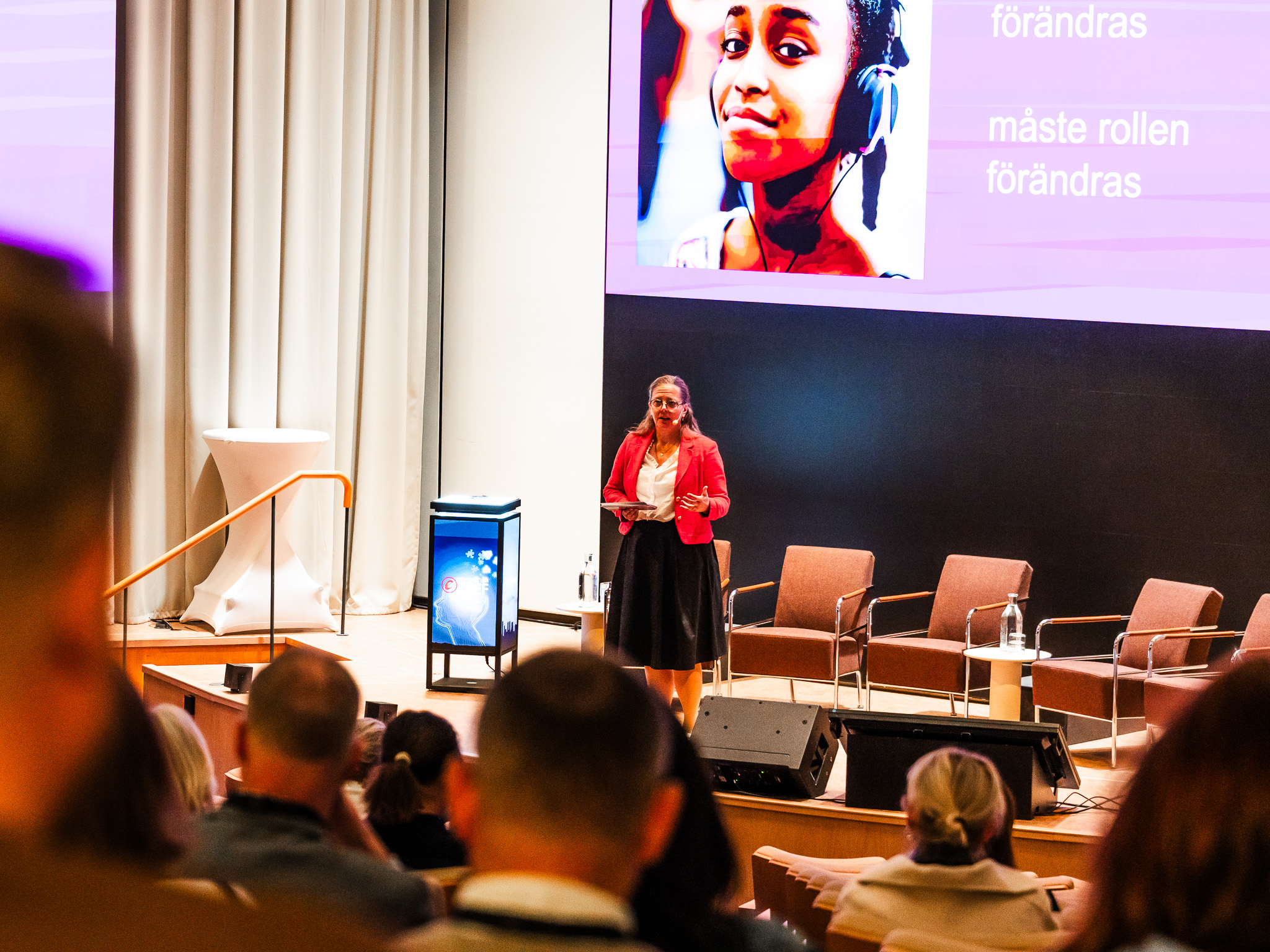 En kvinna i röd kavaj, Åsa Hemingway, står på en stor scen med en presentation bakom sig. På presentationen finns en flicka som ler och har hörlurar på huvudet. I förgrunden syns bakhuvuden från personer i publiken.