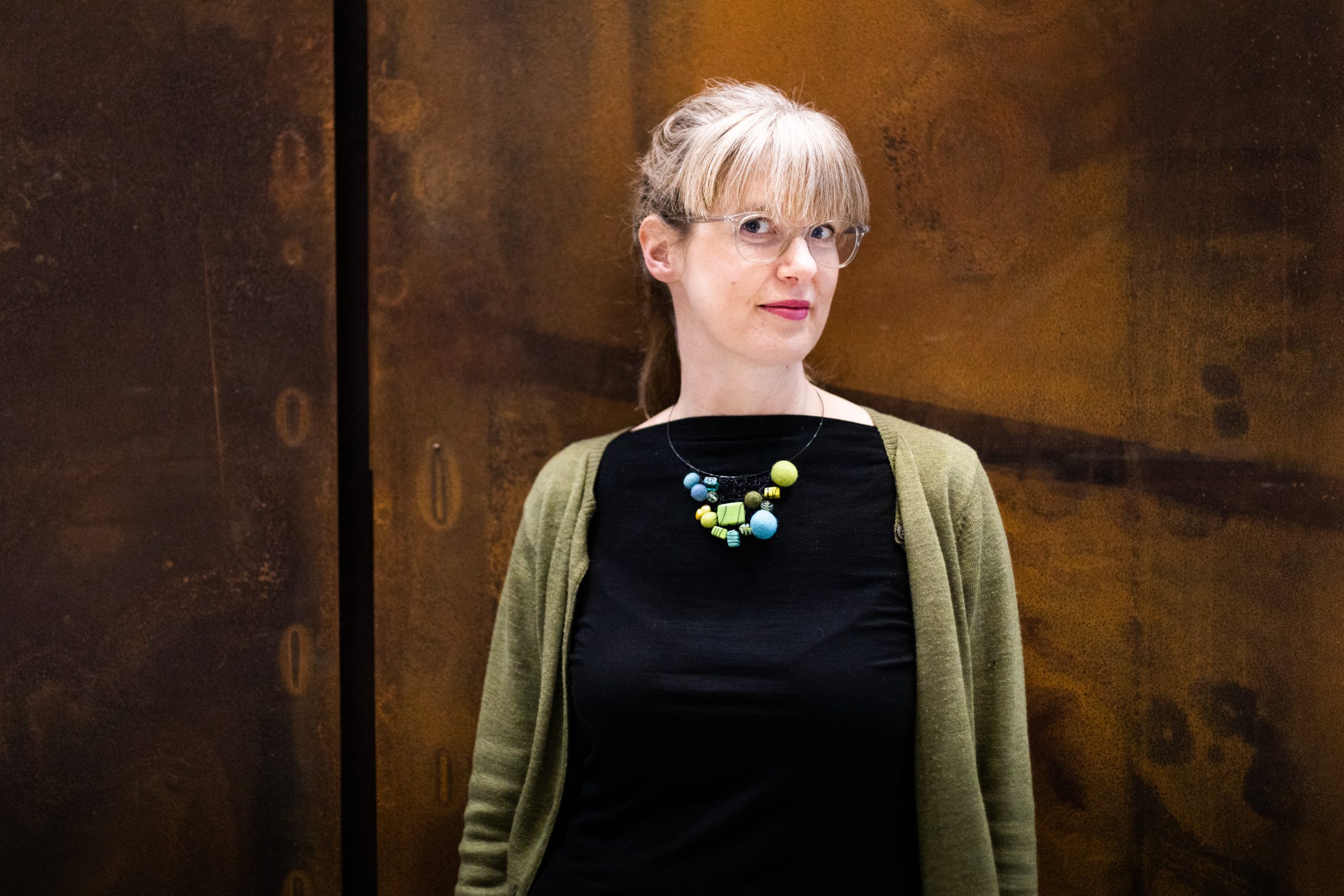 Porträttfoto av MTM:s UX-researcher Hillevi Sundholm som har ett färggrant halsband och en grön kofta och står framför en rostbrun vägg.