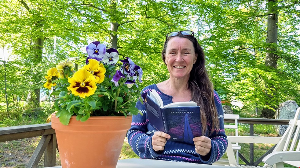 Maria Ståhl sitter utomhus med gröna träd bakom sig och en blomkruka i förgrunden. Hon håller i en blå bok och ler.