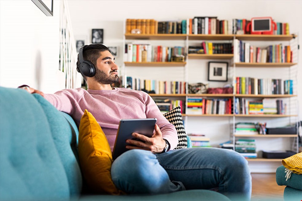 En man sitter i en vardagsrum i en turkosblå soffa och har hörlurar på huvudet och håller en surfplatta i handen. Han kollar ut och bokhyllor syns i bakgrunden.