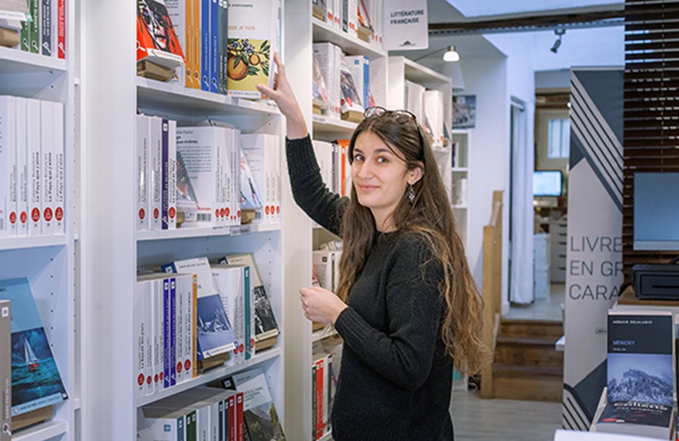 Mélanie  de Carvalho Mélanie  de Carvalho som  arbetar i bokhandeln i Paris, som säljer böcker i storstil. Hon står framför en bokhylla. 
