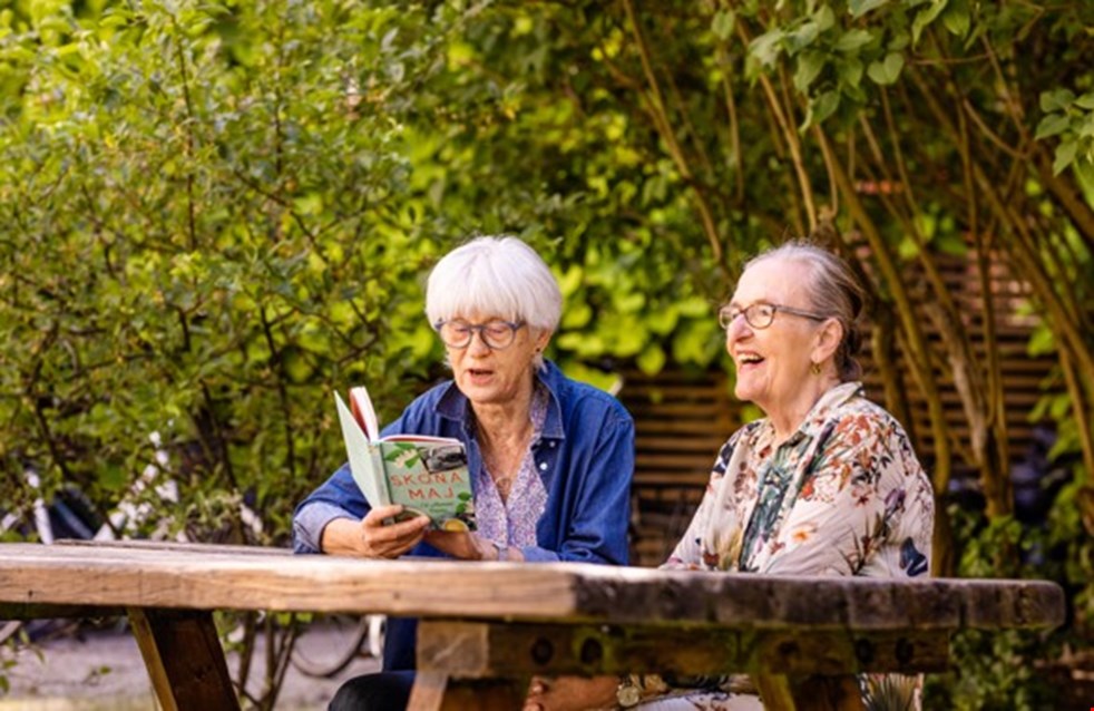 En man och en kvinna sitter i en trädgård. Kvinnan läser ur en bok för mannen.