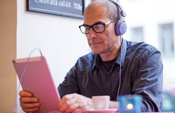 En man sitter på ett café med en läsplatta i händerna och läser en taltidning. Han bär svarta glasägen och en grå skjorta med en grå tröja under. På huvudet har han ett par lila hörlurar som är kopplade till en läsplattan.