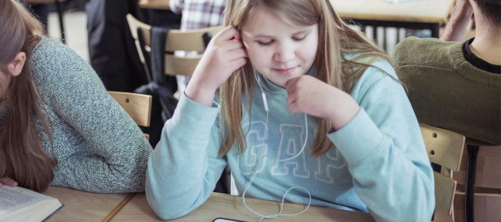 BIlden visar en tjej i ljusblå hoodtröja sittandes vid ett bord i klassrummet med hörlurar i öronen och kollar ner på en läsplatta.
