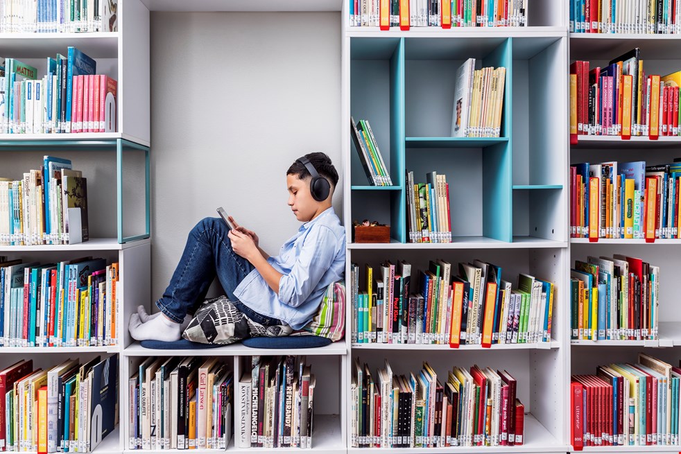 En pojke sitter i en hylla i ett bibliotek, omgiven av böcker och lyssnar på något i hörlurar samtidigt som han tittar på en skärm.