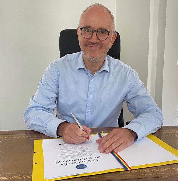 MTM:s generaldirektör Magnus Larsson skriver på Deklarationen för en stark demokrati. Han sitter vid ett skrivbord med en penna i handen och ler in i kameran.
