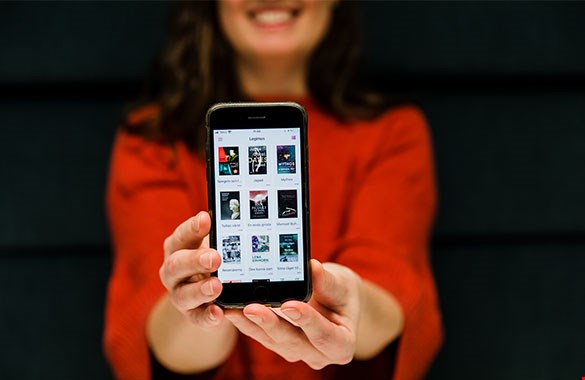 Bilden visar en kvinna i en röd tröja och långt brunt hår där endast halva ansiktet syns. Bakom henne syns en mörk vägg och framför sig håller hon upp en smarttelefon där nya Legimusappen syns på skärmen.
