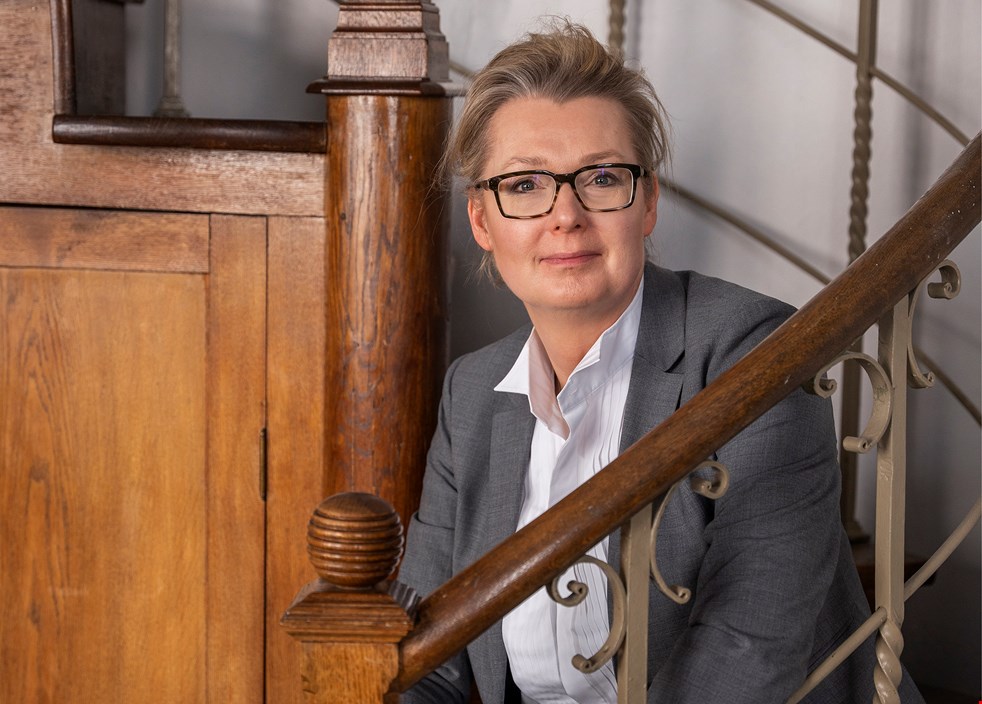 Skolminister Lina Axelsson Kihlblom sitter i en äldre trätrappa i ett bibliotek. Hon har mellanblont uppsatt hår, glasögon, grå kavaj och vit skjorta.