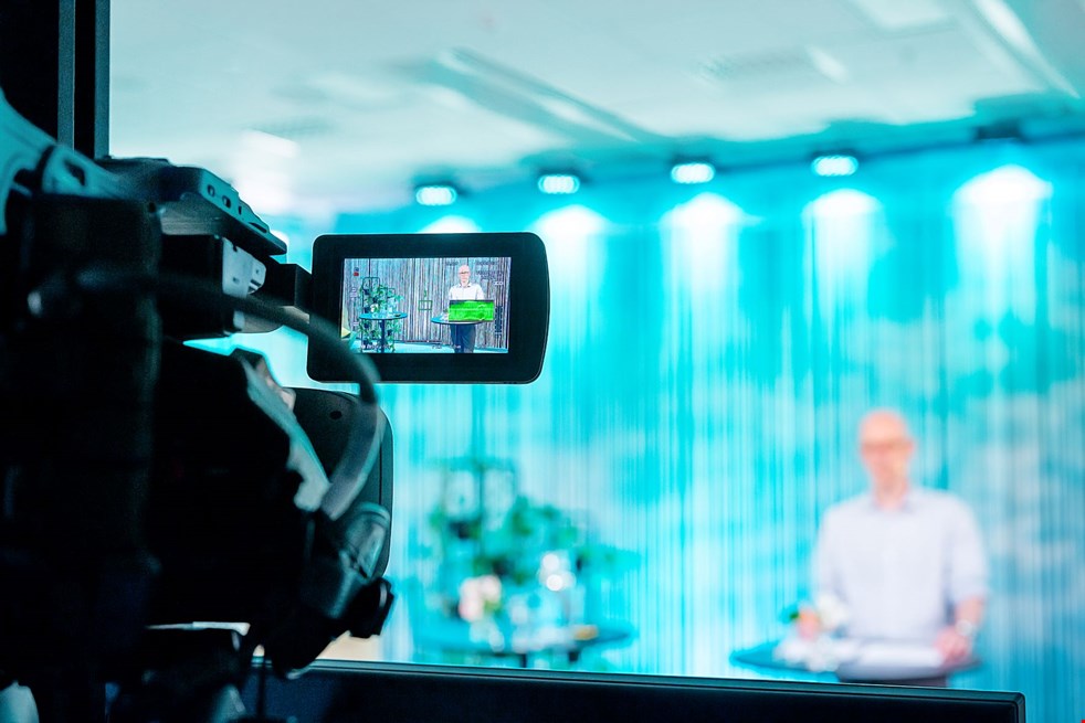 En videokamera filmar en person som står i en turkosblå sändningsstudio. Skärpan ligger på kameran och personen på scen är inte identifierad.