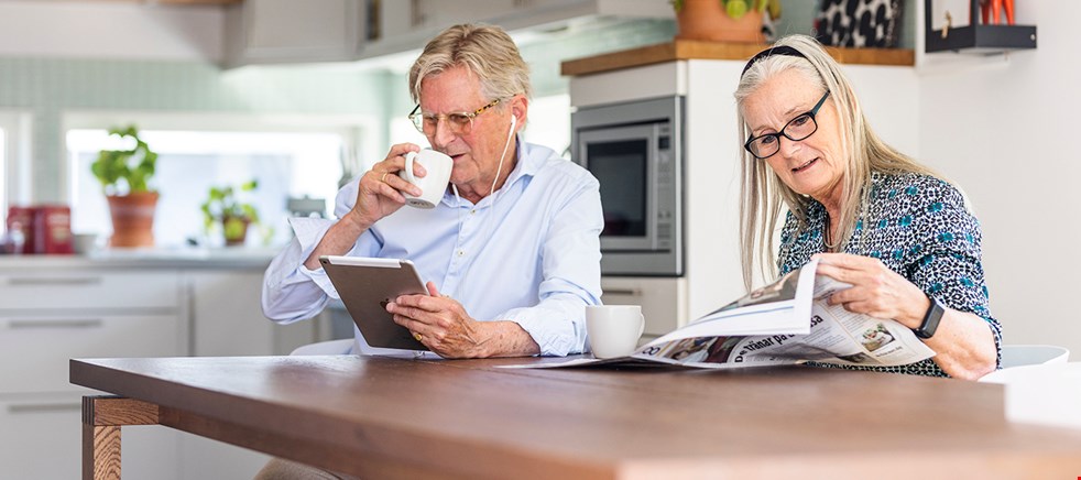 Ett äldre par sitter i ett kök och läser tillsammans. Mannen läser taltidningen på en läsplatta med lurar på huvudet och kvinnan läser papperstidningen 8 Sidor.