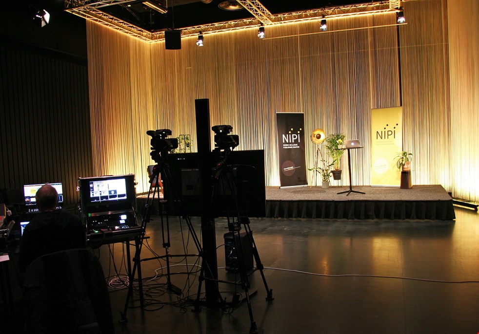 Ett fotografi från förra NIPIkonferensen 2020. Scenen är upplyst med lampor som ger ett guldaktigt sken och på scenen står roll-ups med NIPIs logotyp.