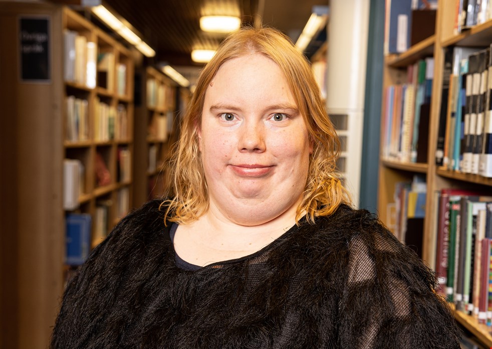 Maila Goman står i ett bibliotek med bokhyllor på båda sidorna om sig. Hon ler och tittar in i kameran.