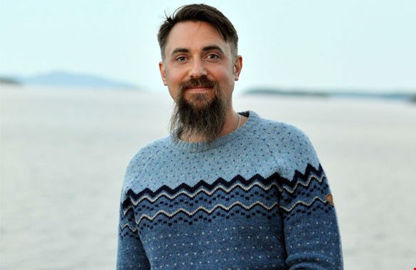 Bilden visar Sebastian Åkesson i en blåstickad tröja. Han har mörkt hår och långt skägg. Suddigt i bakgrunden syns ett berg.